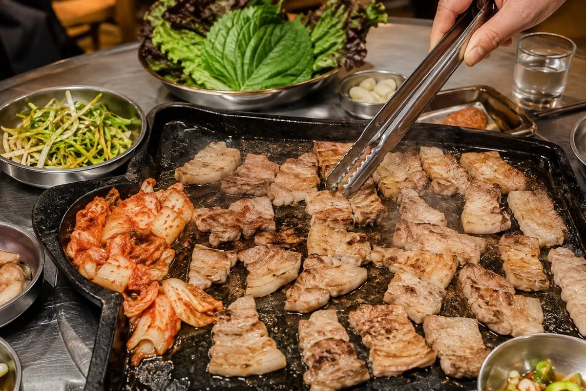 AI concept: pork belly and kimchi cooking on a black table grill, tongs turning one piece, ssam greens and banchan around it.